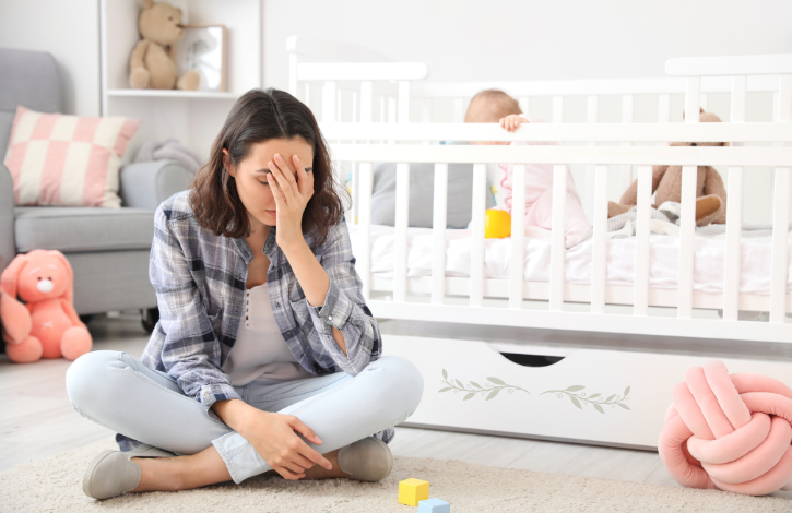 Woman holding her head in her hands, sitting beside a crib with a baby. 